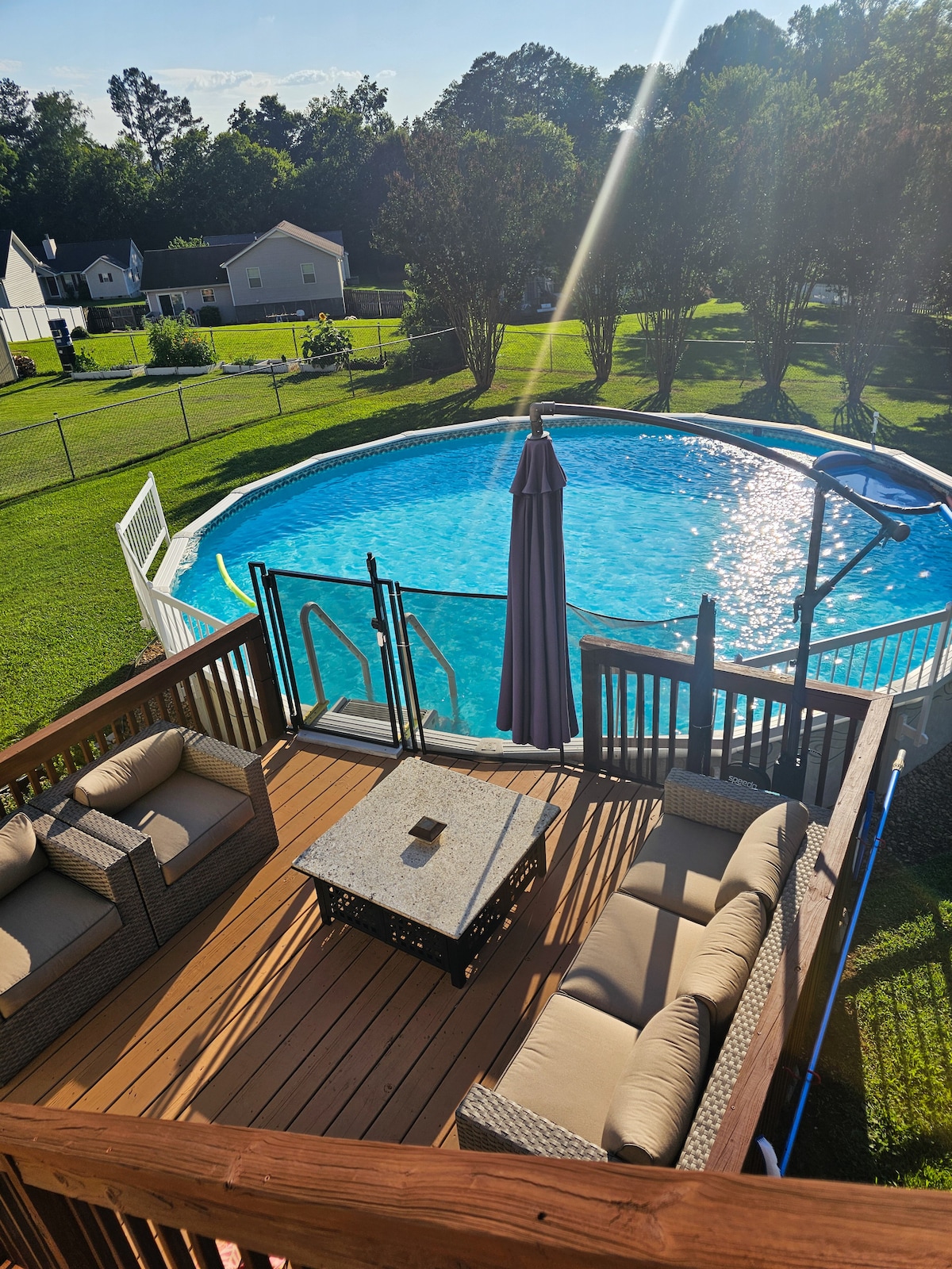 An upper deck area is furnished with a seating arrangement and a table, offering a serene view of the spacious backyard. A large above-ground pool with a ladder is visible, surrounded by a fence and lush green grass, creating a relaxing outdoor space.