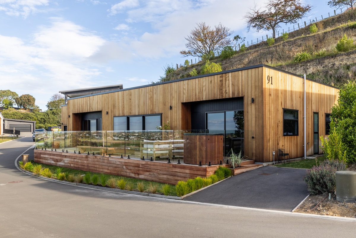 A contemporary wooden structure is set against a backdrop of rolling hills. The building features large glass windows and a fenced patio area with a low wooden railing. Lush greenery and planters line the entrance, and a paved driveway leads up to the property.