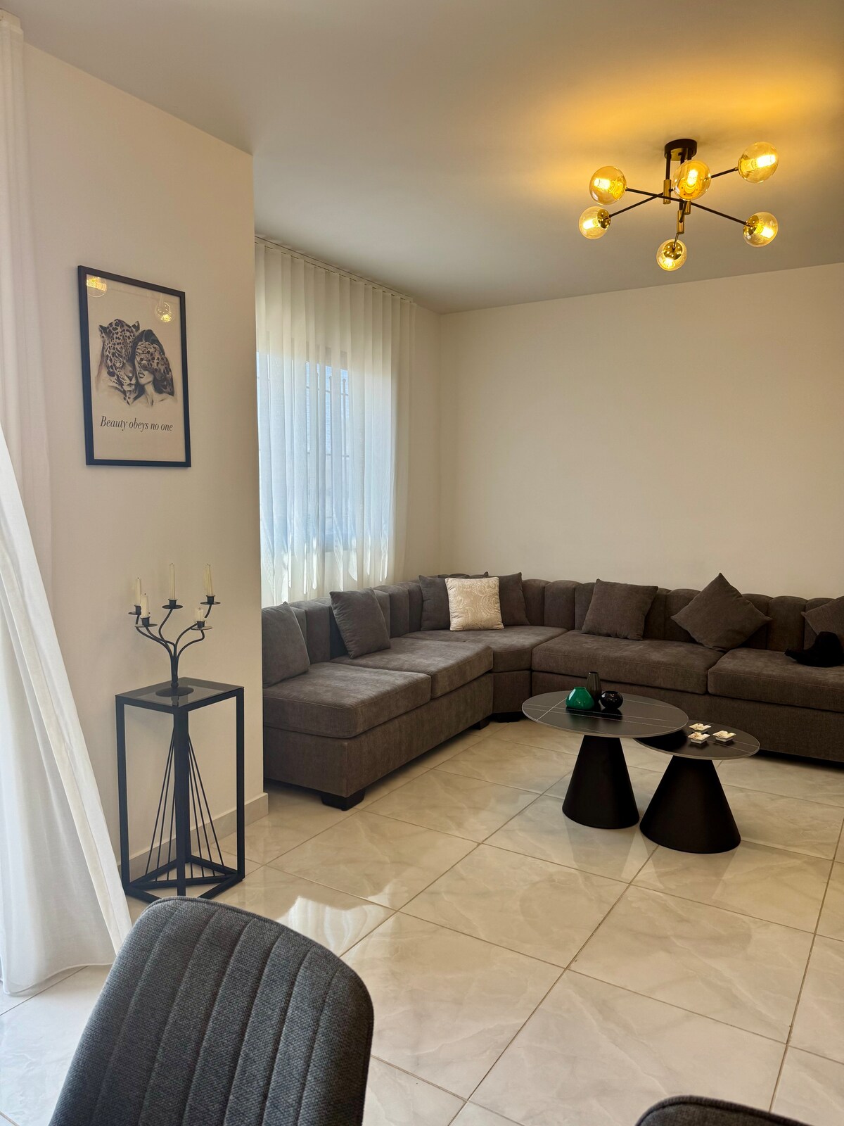 A modern living area features a sectional sofa adorned with decorative pillows, complemented by a pair of black side tables. Natural light filters through sheer curtains, illuminating the glossy tiled floor. A minimalistic floor lamp and wall art add subtle interest to the space.