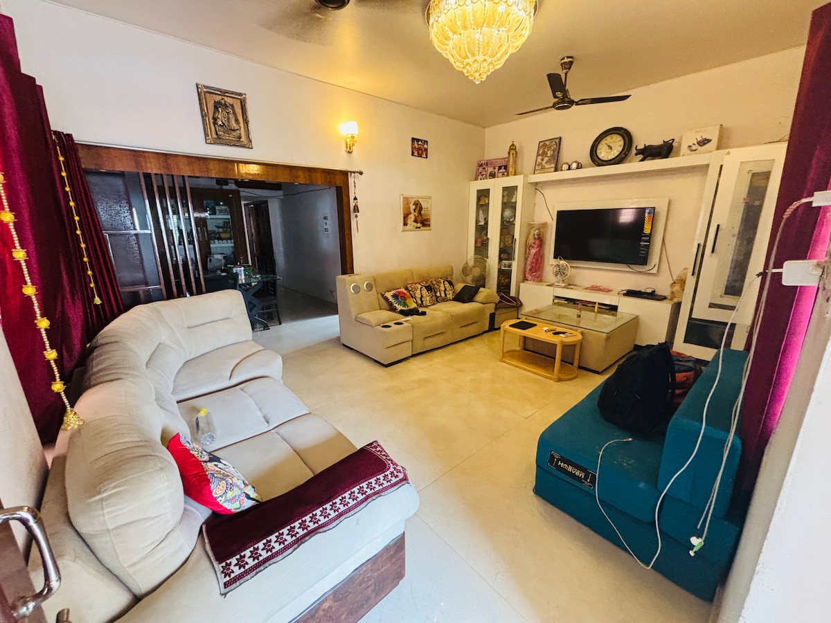 A comfortable living area is furnished with a sectional couch, a two-seater sofa, and a coffee table. A large television is mounted on the wall, while decorative wall art and a ceiling fan contribute to the room's ambiance. Light streams in through windows, complemented by red curtains.