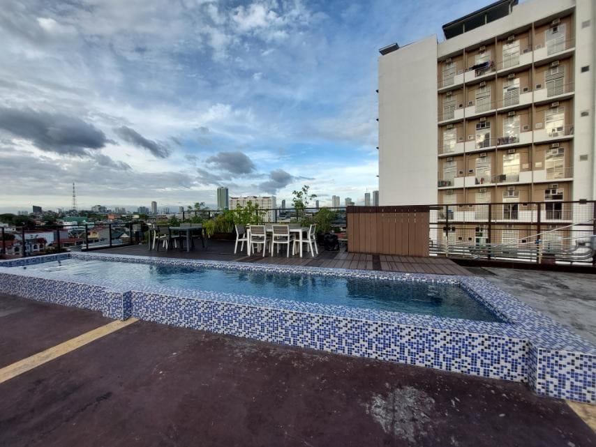 An outdoor rooftop area features a small swimming pool with blue mosaic tiles, surrounded by a wooden deck. Several white tables and chairs are positioned nearby, offering a space to relax. The skyline of the city can be seen in the background under a partly cloudy sky.