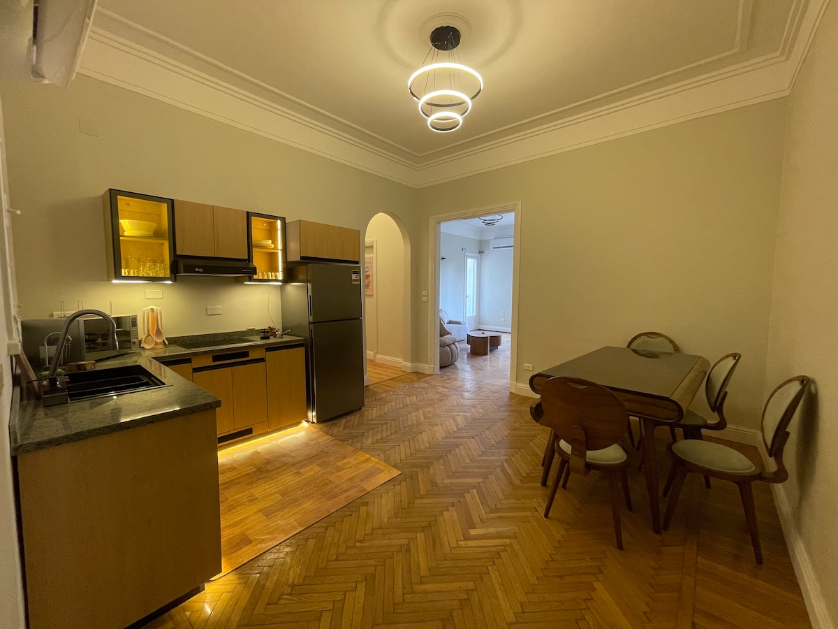 An open-layout kitchen and dining area is presented, featuring sleek cabinetry and modern appliances, including a refrigerator and stove. A dining table with chairs is situated adjacent to the kitchen, while light fixtures enhance the room's ambiance. The space is designed in a warm, inviting style.