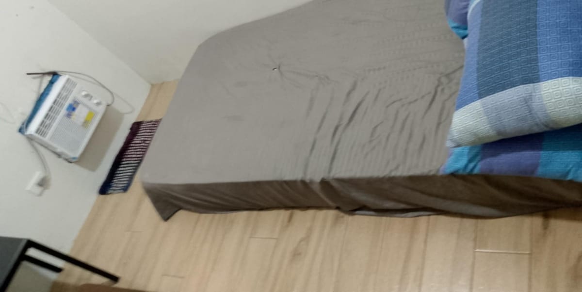 A spacious bed with a neutral-toned cover is positioned on a light wood floor. Pillows with various blue patterns are placed at the head of the bed. An air conditioning unit is mounted on the wall next to the sleeping area.