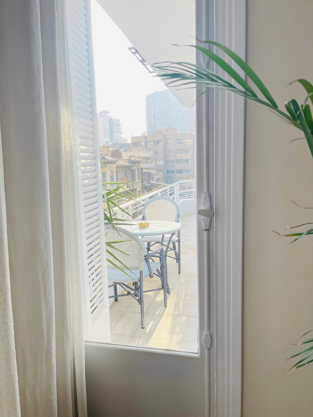 A view is presented through a partially opened window. A small outdoor balcony is visible, furnished with a table and two chairs. Light filters in, highlighting the greenery from a nearby plant. City buildings are seen in the background, adding to the urban ambiance.