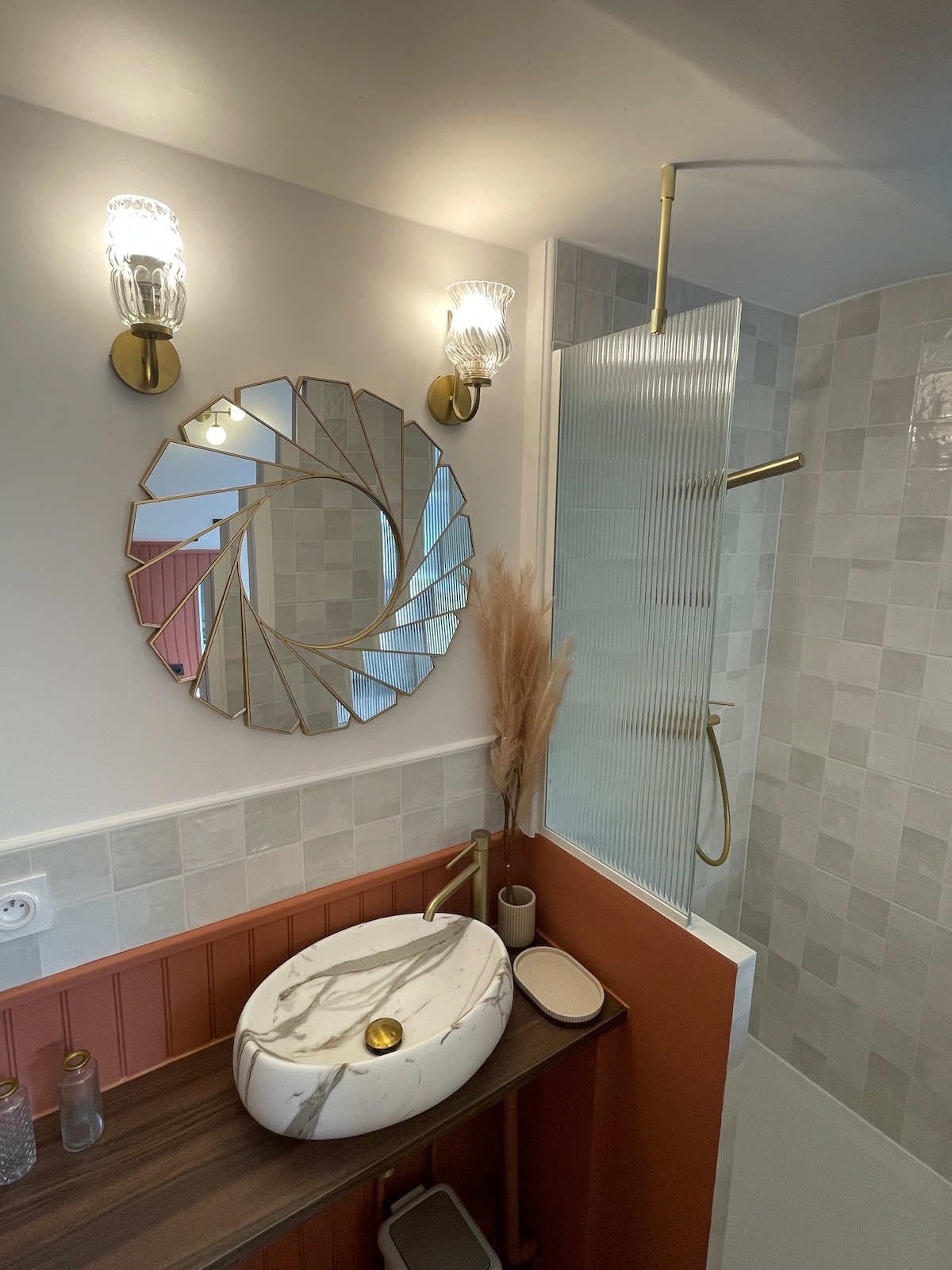 A modern bathroom is depicted, featuring a round, stylish mirror above a marble sink set on a wooden surface. A tall, glass shower with brass fixtures is visible, along with elegant wall sconces providing soft light. The tiled walls display a mix of textures and colors.