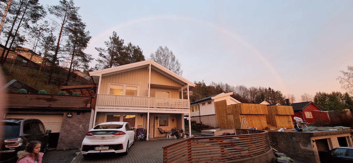 A two-story house is showcased with a light-colored facade and a covered balcony. The surrounding area features a mix of greenery and neighboring homes, with a wooden fence partially visible. A rainbow arcs across the sky, adding a colorful backdrop to the scene.