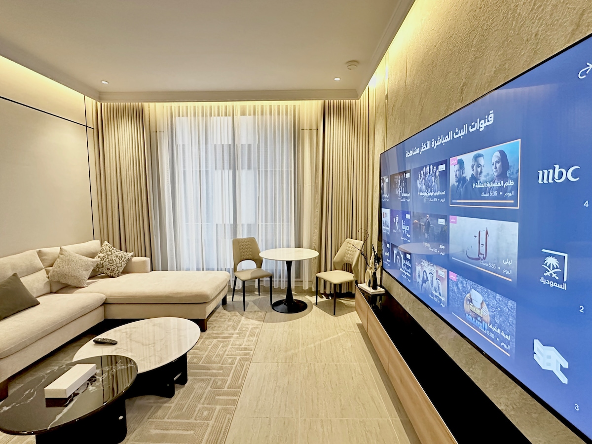 A modern living area features a spacious cream-colored sofa, complemented by subtle decorative pillows. A sleek coffee table sits in front, with a small dining table and chairs nearby. A large screen displays various entertainment options, while natural light filters through the floor-to-ceiling curtains.