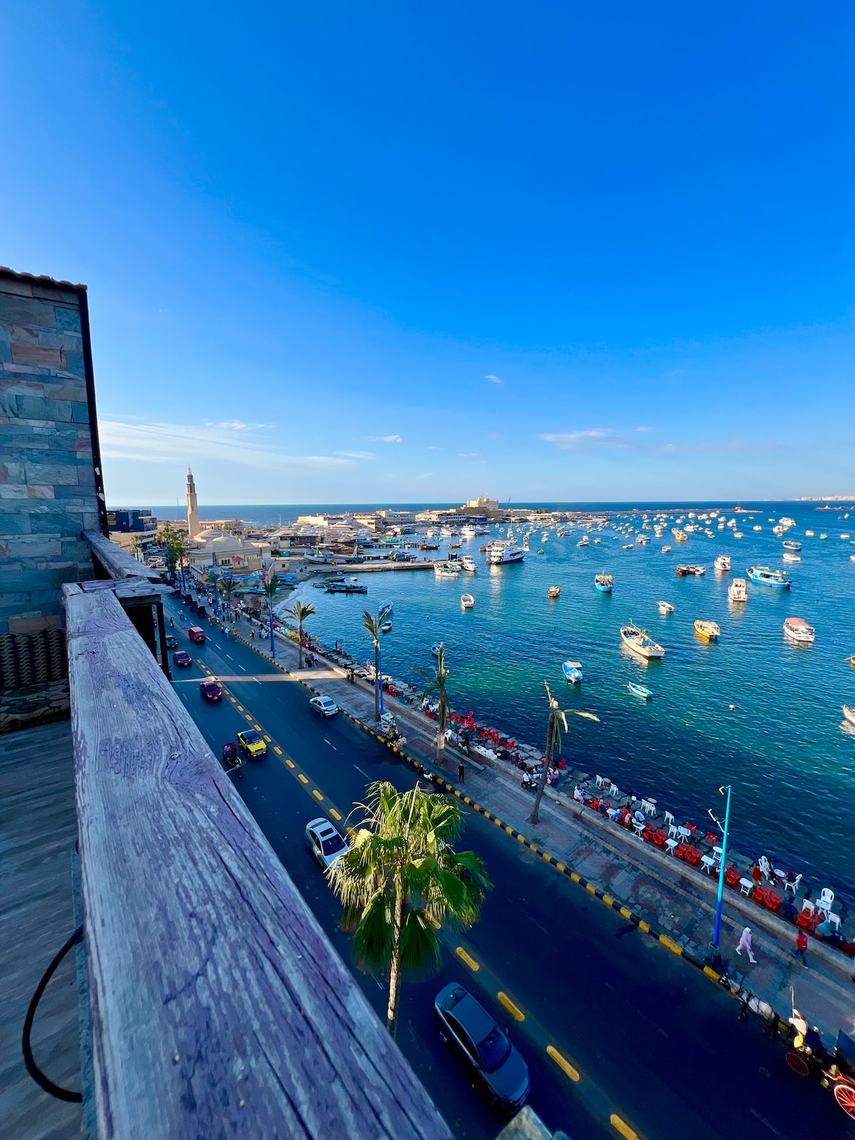 A panoramic view from the balcony reveals the Mediterranean Sea, dotted with numerous colorful boats. A vibrant waterfront road stretches along the coast, lined with palm trees. The sky appears clear and blue, enhancing the overall tranquil seaside atmosphere.