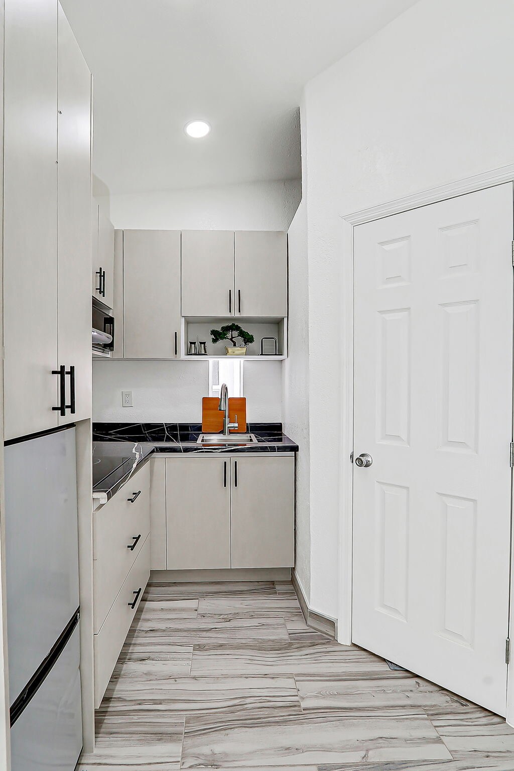 A compact kitchen area features light-colored cabinetry and a modern stainless-steel refrigerator. Counter space is lined with dark countertops, while appliances include a microwave and stove. A door is visible on the right, leading to another area, and the floor has a wood-like tile finish.