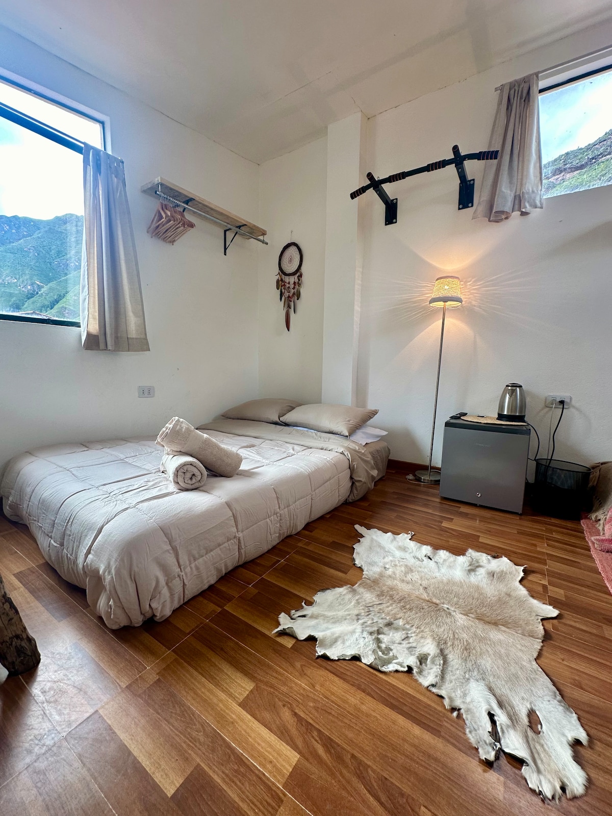A compact sleeping area is depicted, featuring a low bed covered with soft linens and plush pillows. Nearby, a small refrigerator and kettle are visible. Natural light enters through two windows, creating a bright atmosphere. A decorative rug lies on the wooden floor.
