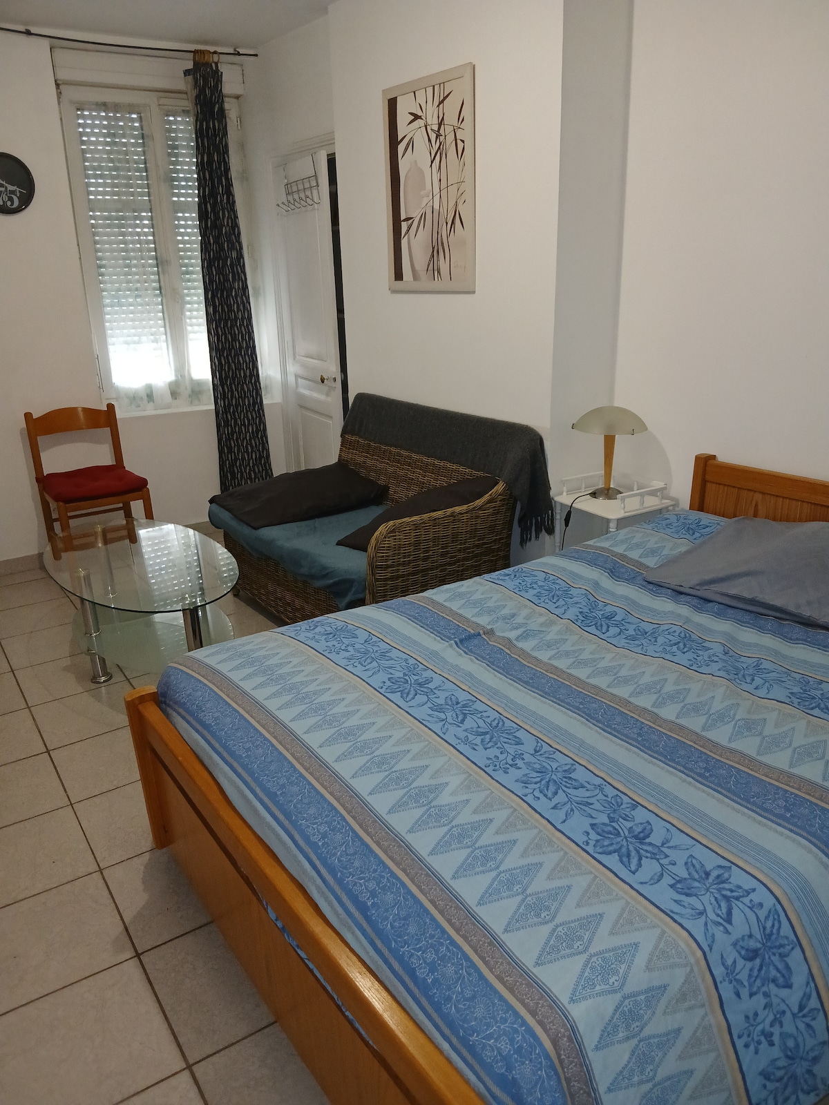 A comfortable sleeping area includes a spacious bed with a patterned blue and white coverlet. A small glass table is positioned next to a cozy seating area featuring a cushioned chair and a wicker loveseat. Natural light enters through the window, partially covered by light curtains.