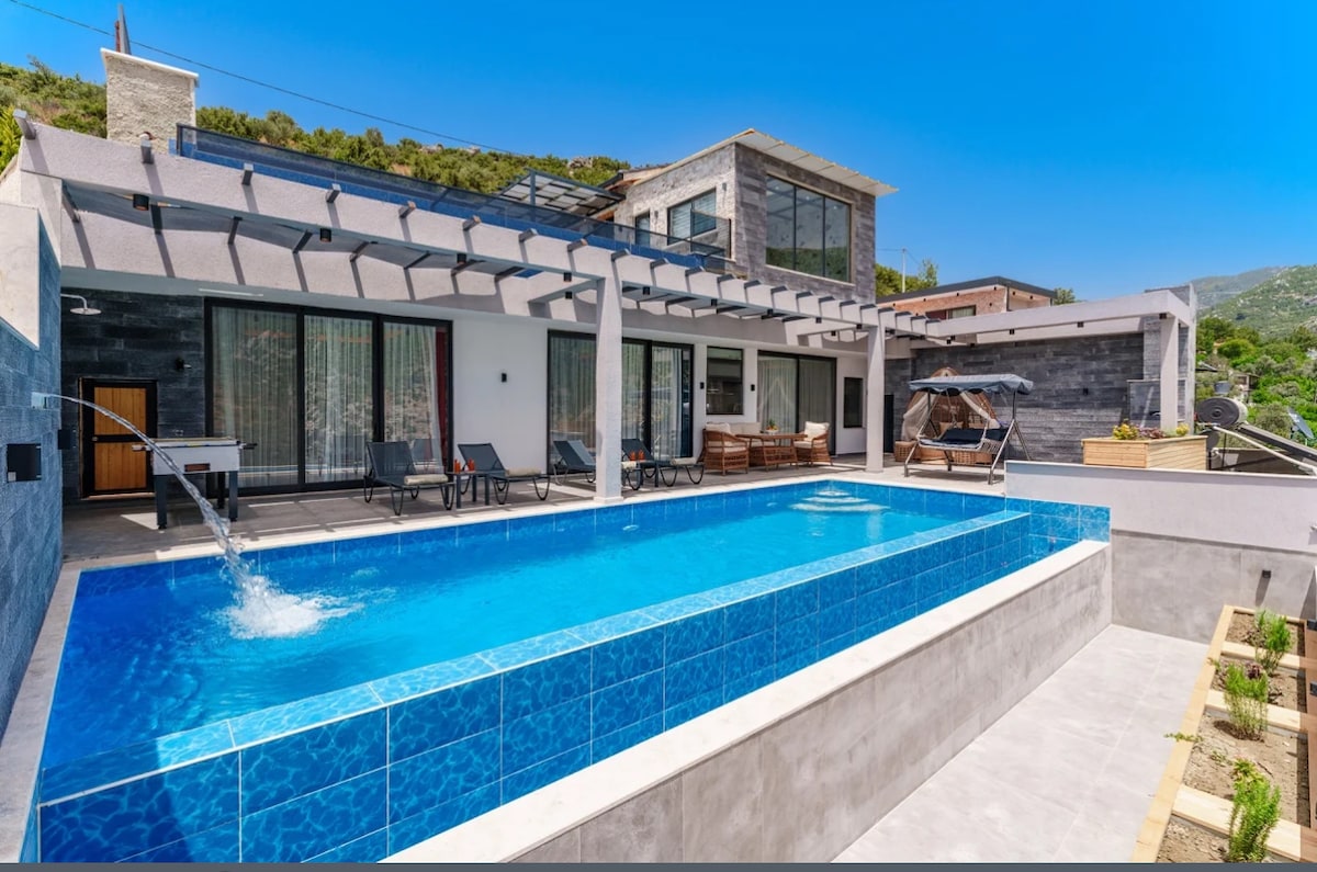 A bright outdoor area features a swimming pool surrounded by modern lounge chairs under a pergola. An outdoor dining set is visible nearby, along with a shaded resting spot. The backdrop showcases lush greenery against a clear blue sky.