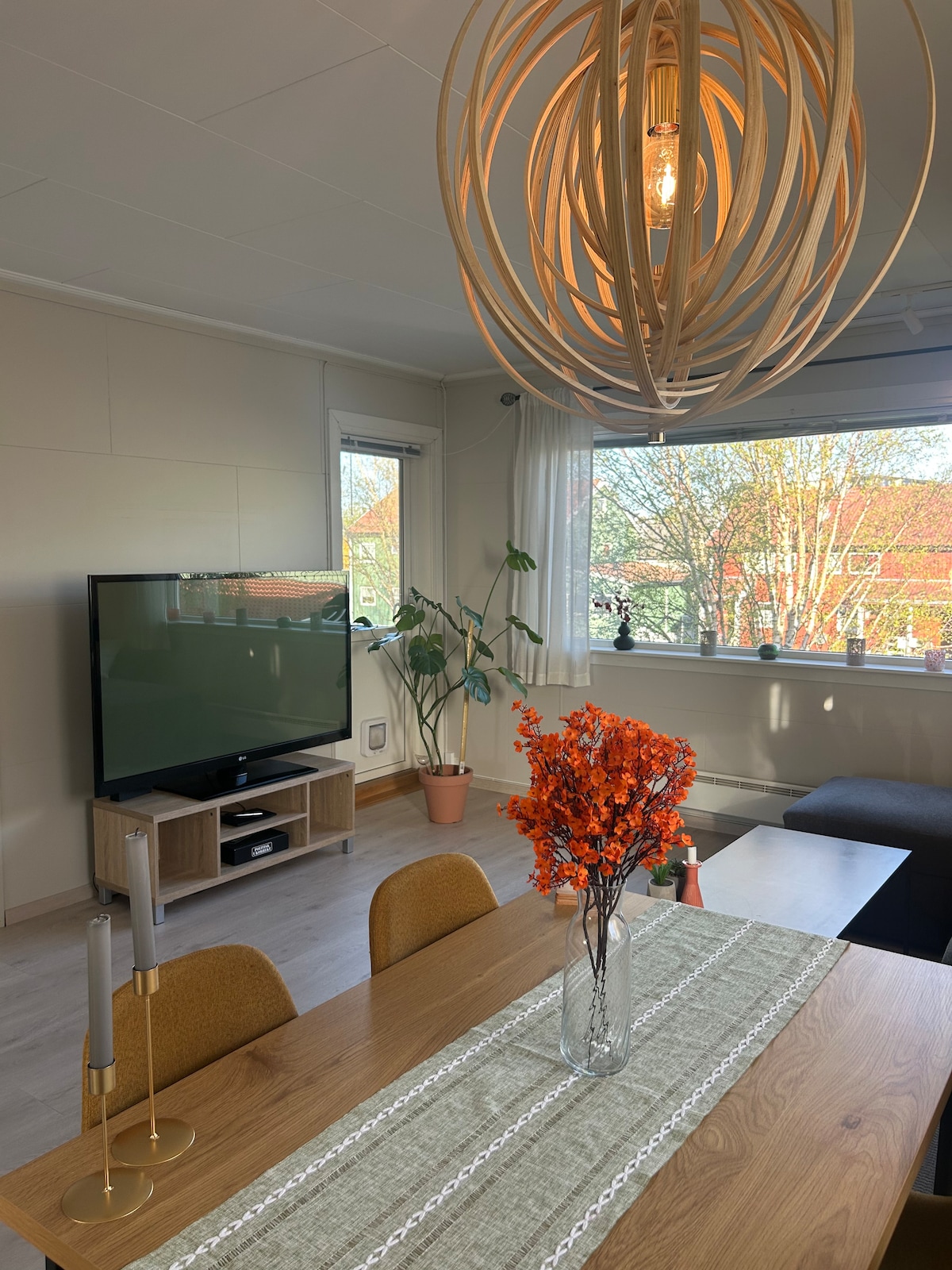 A dining area features a wooden table with two chairs and a decorative runner. A bouquet of orange flowers sits in a vase on the table. A cozy seating area with a dark sofa is adjacent to a TV stand, illuminated by a unique pendant light overhead.