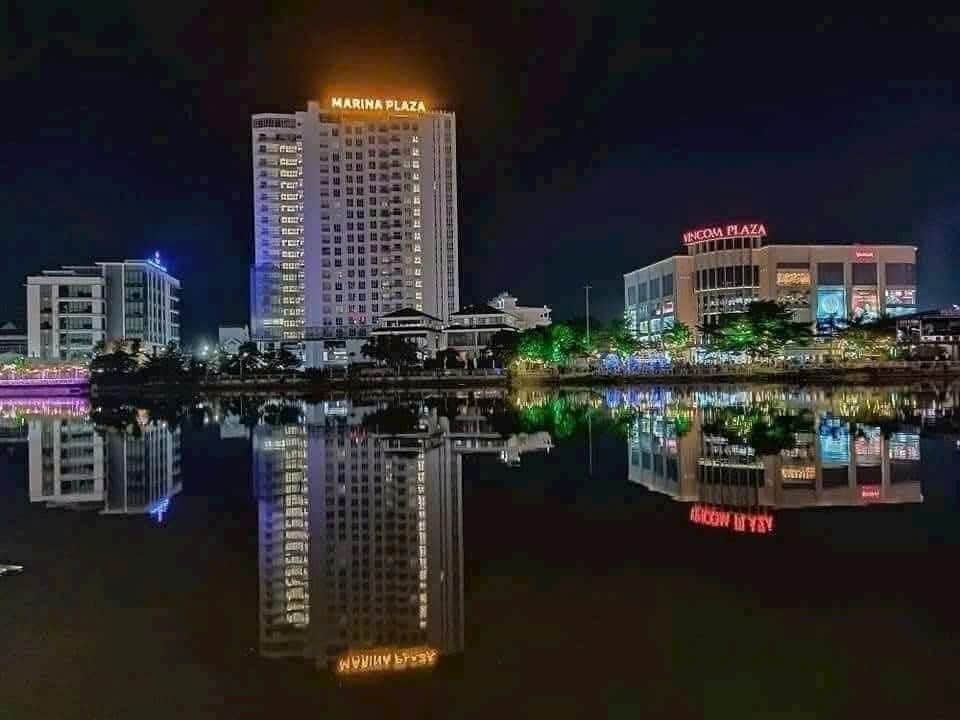 A tall building named Marina Plaza is illuminated at night, showcasing its reflection in calm waters below. Nearby structures are also faintly visible along the waterfront, contributing to the urban skyline of Long Xuyen city with colorful lights glimmering in the water.