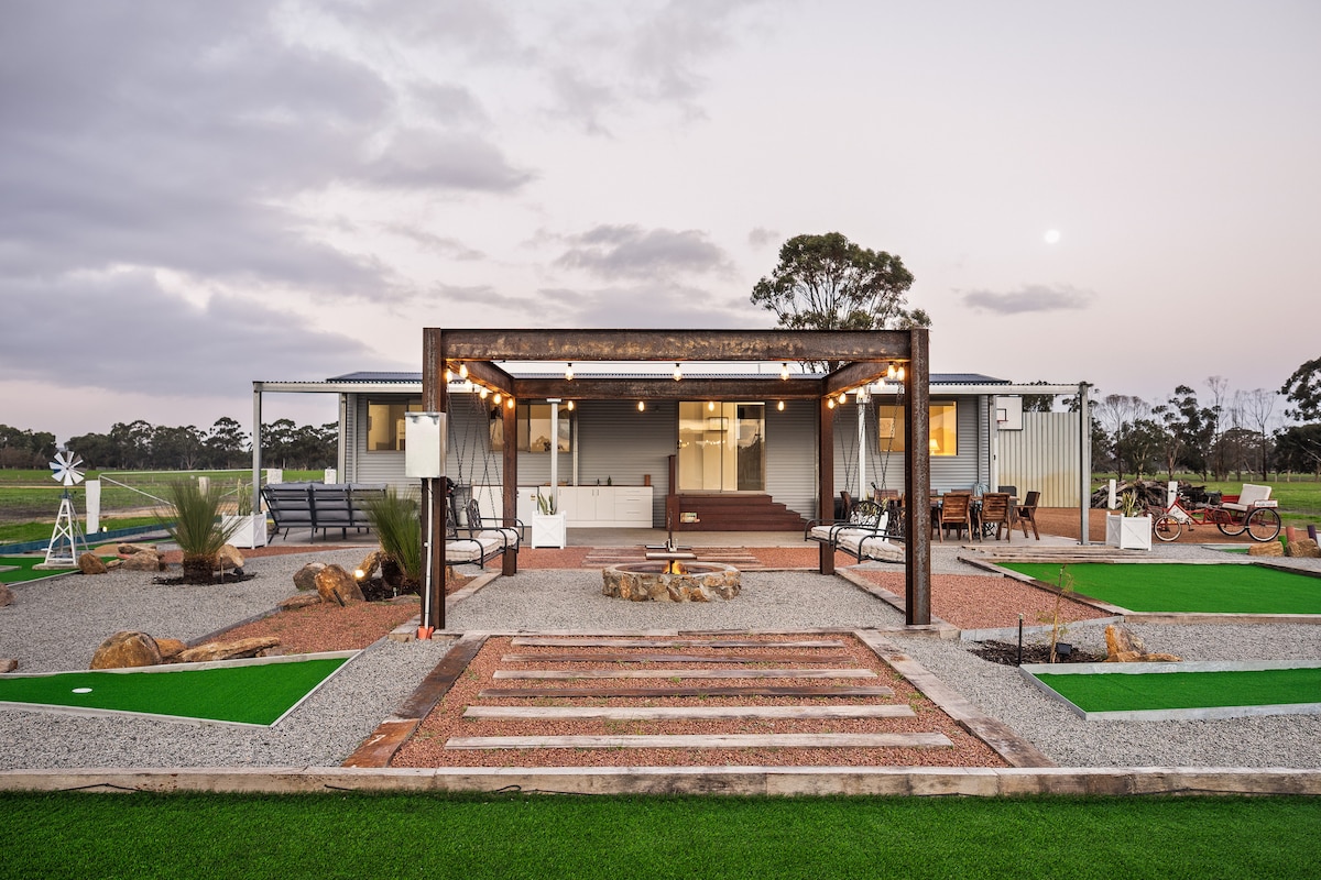 The cabin's exterior showcases a spacious patio area, framed by wooden beams and string lights. Pathways lead towards the entrance, with landscaped greenery and a mini golf course visible in the foreground. Natural stone accents are positioned around the space, enhancing the rural setting.