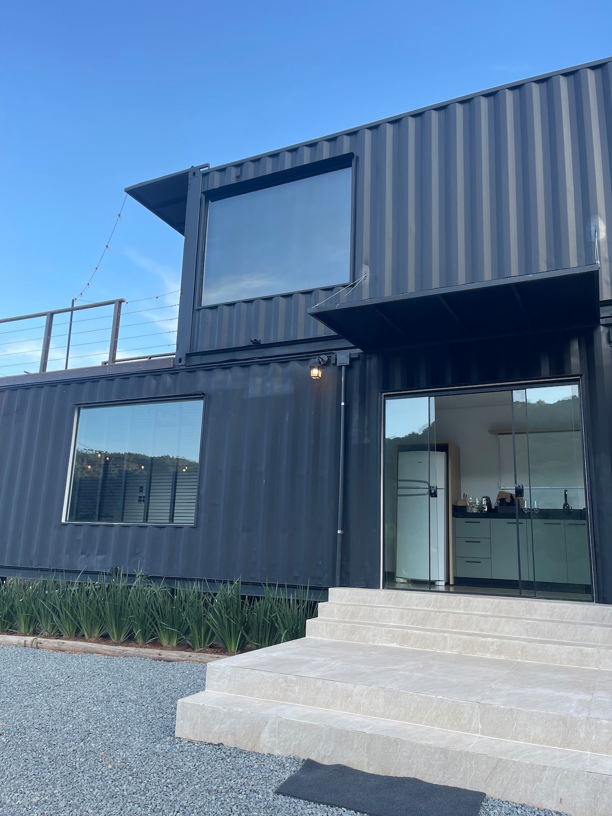 A modern two-story structure features a sleek black exterior made of shipping containers. Large windows allow natural light to fill the interior. A stone staircase leads to the entrance, with gravel and greenery surrounding the base of the building.