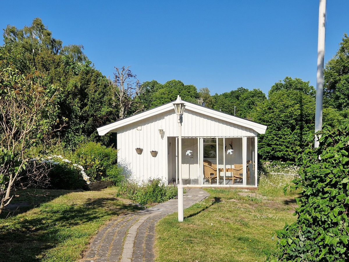 A charming white house is set amidst lush greenery, featuring a pathway leading to its entrance. Large glass doors provide a view of the inviting interior space, while flowerbeds and greenery surround the exterior, creating a serene and natural environment.