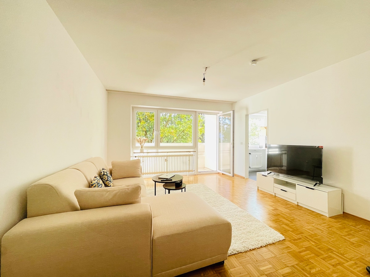 A bright living area is featured, showcasing a light-colored sectional sofa with decorative pillows. A coffee table is positioned on a soft, plush rug, while large glass doors open to a balcony. Natural light fills the room, which includes a modern television on a white entertainment unit.