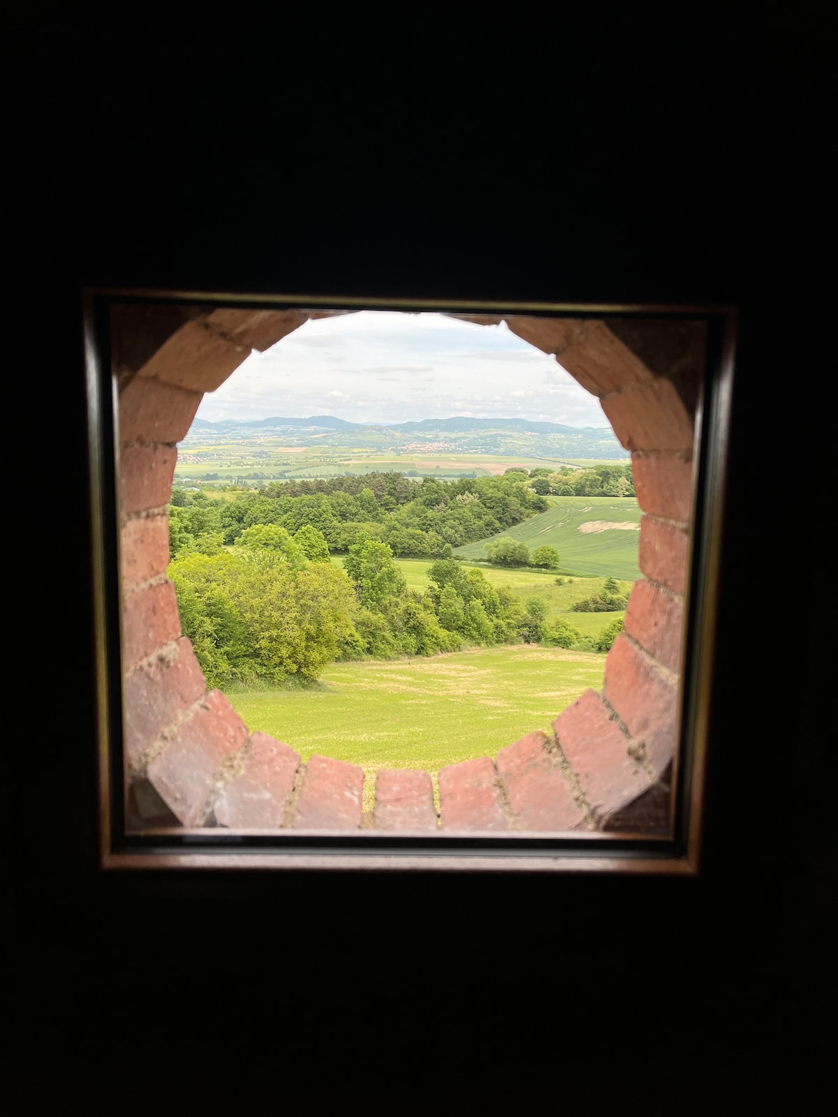 A circular window framed by rustic bricks opens to a lush landscape. Gentle hills and verdant fields extend into the distance, under a sky scattered with clouds. The natural scenery offers a serene view, reinforcing the retreat's tranquil setting.
