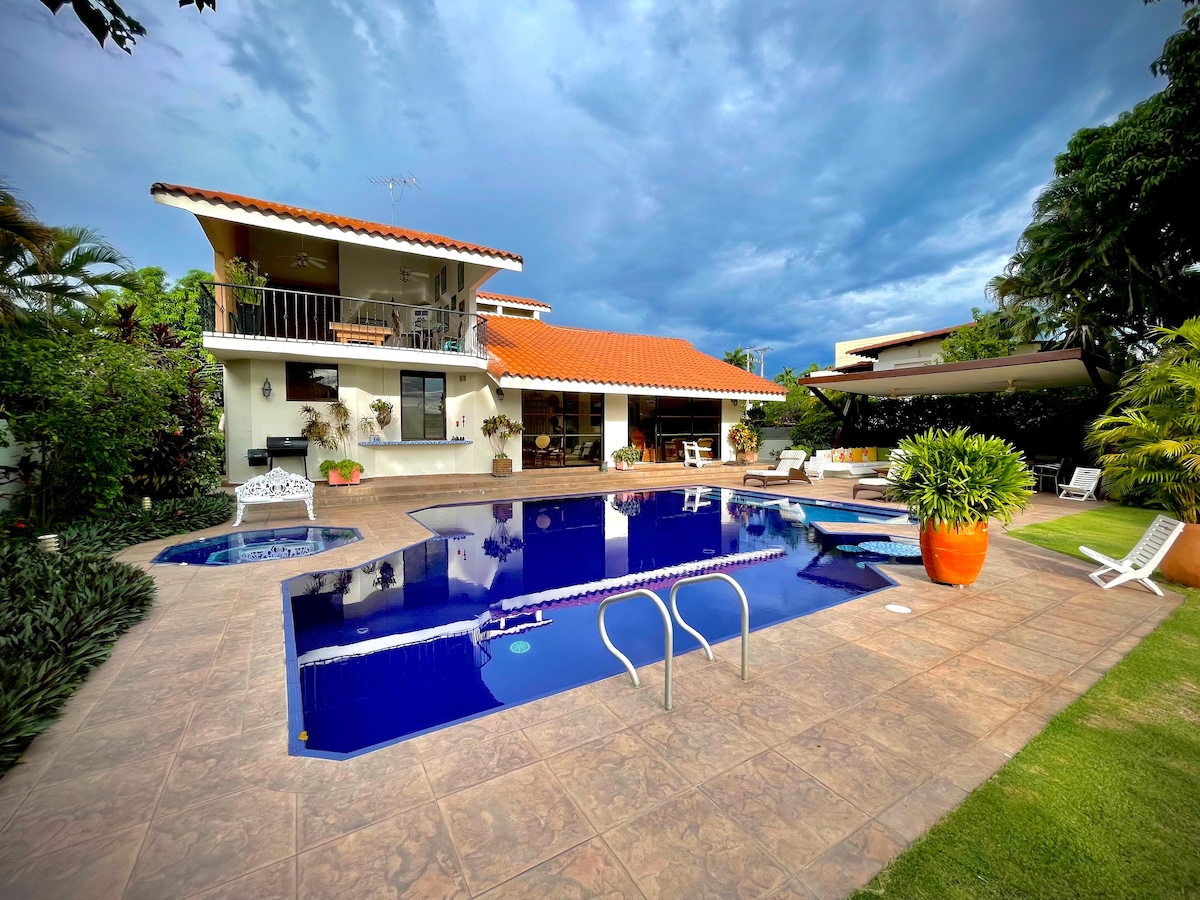 A spacious outdoor area is highlighted by a large, recently renovated pool surrounded by a sunbathing deck. The house features an orange-tiled roof and multiple stories, with lush greenery and potted plants enhancing the serene environment.