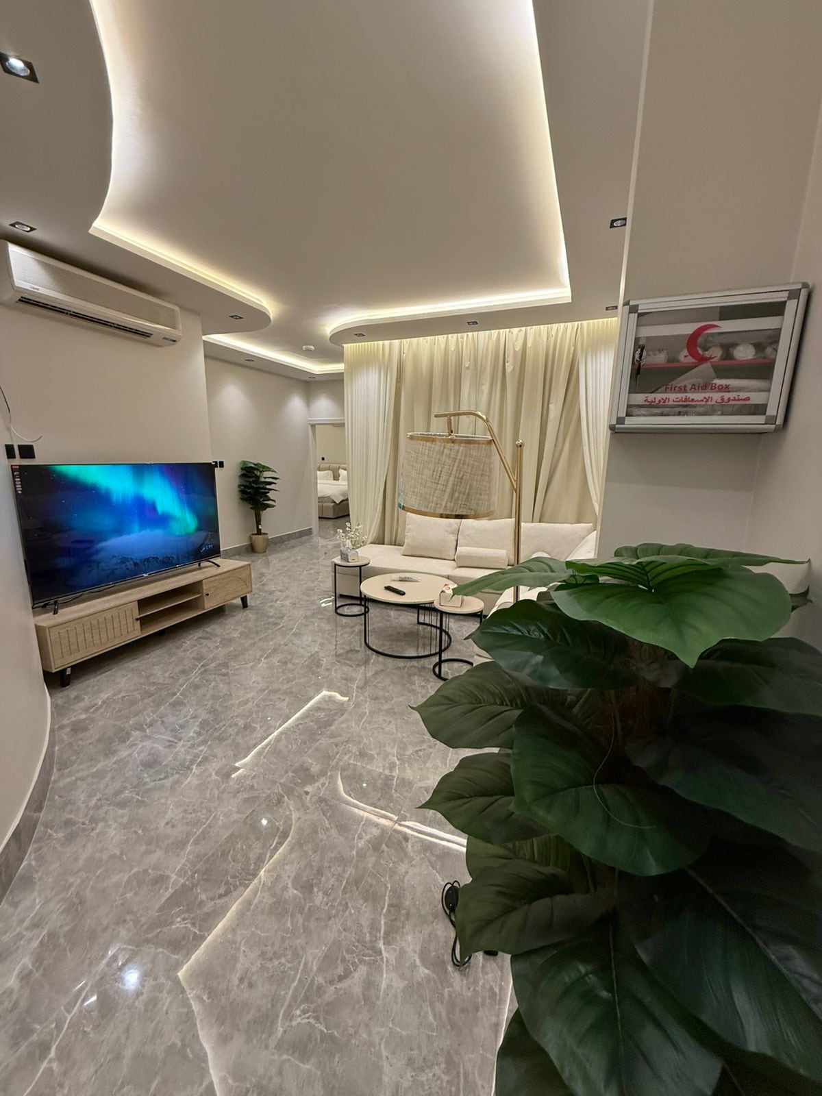 A modern living area is featured with a sleek linear design. A large television is mounted on the wall opposite a comfortable seating area with a light-colored sofa and coffee tables. Soft lighting enhances the ambiance, while decorative indoor plants add a touch of greenery.