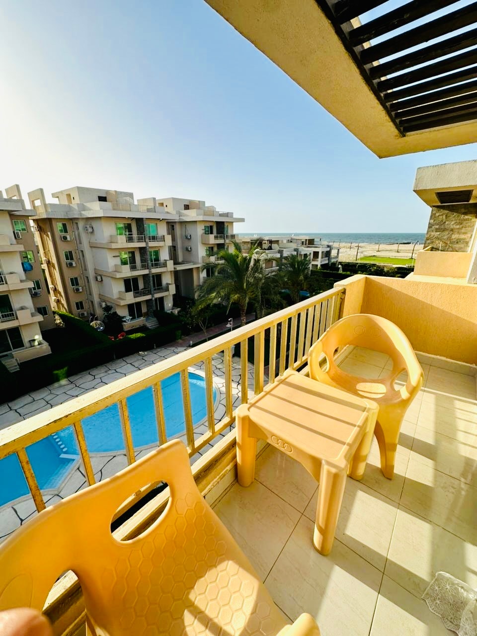 A balcony is depicted, featuring two yellow plastic chairs and a small side table. A clear view of a pool and surrounding greenery is visible below, with the ocean in the distance. Bright sunlight enhances the openness of the space.