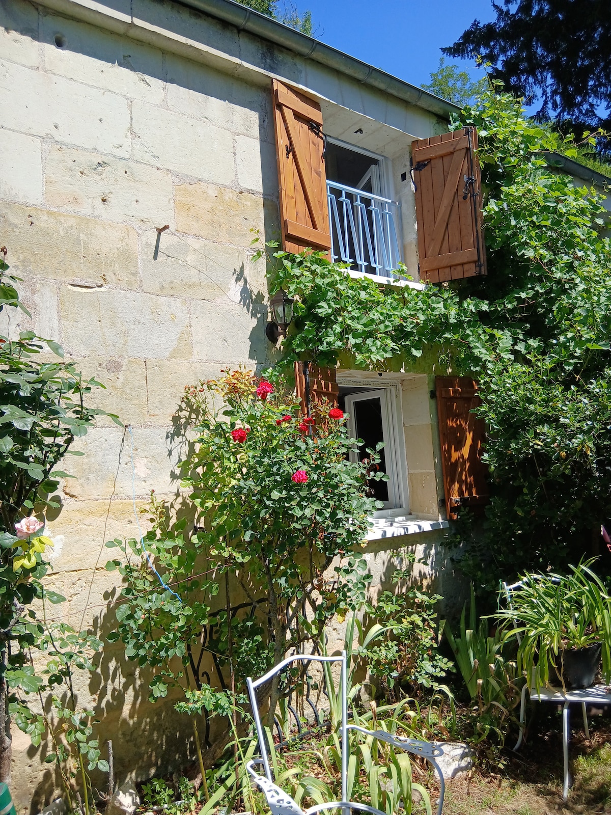The exterior of a rustic two-story house is displayed, featuring stone walls adorned with climbing plants. Bright red flowers flourish near the window, which is framed by wooden shutters. Sunlight bathes the scene, highlighting the greenery and a simple white chair nearby.