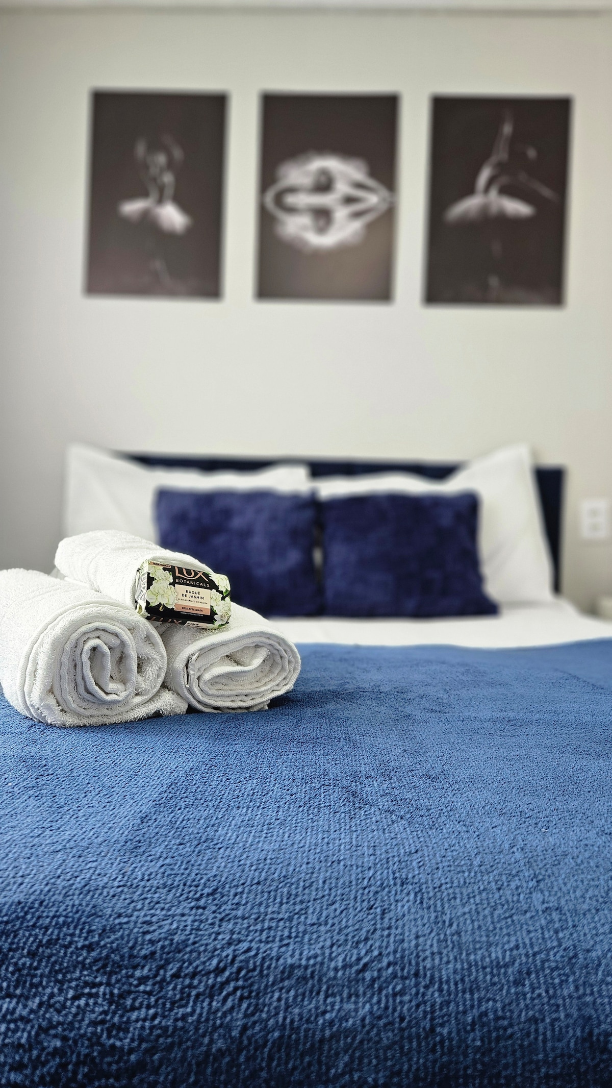 A neatly arranged bed is covered with a textured blue blanket. Plush white towels are rolled and placed at the foot of the bed. Above the bed, three framed black-and-white photographs featuring ballet dancers are displayed on the wall.