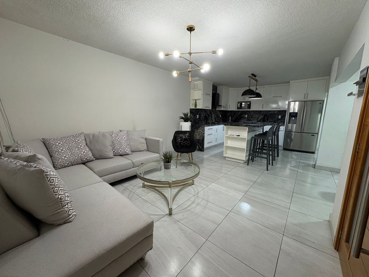 A modern living space is shown, featuring a light grey sectional sofa adorned with decorative pillows. A glass coffee table sits on polished tile flooring, with a small plant placed on it. The open kitchen area, equipped with dark countertops and white cabinetry, is visible in the background.