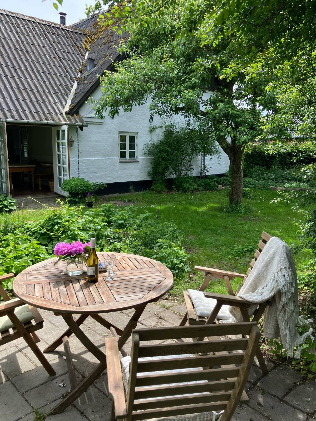 An outdoor seating area features a round wooden table surrounded by folding chairs. A small vase with pink flowers sits on the table. Lush greenery surrounds the space, with a view of the white house wall partially visible in the background.