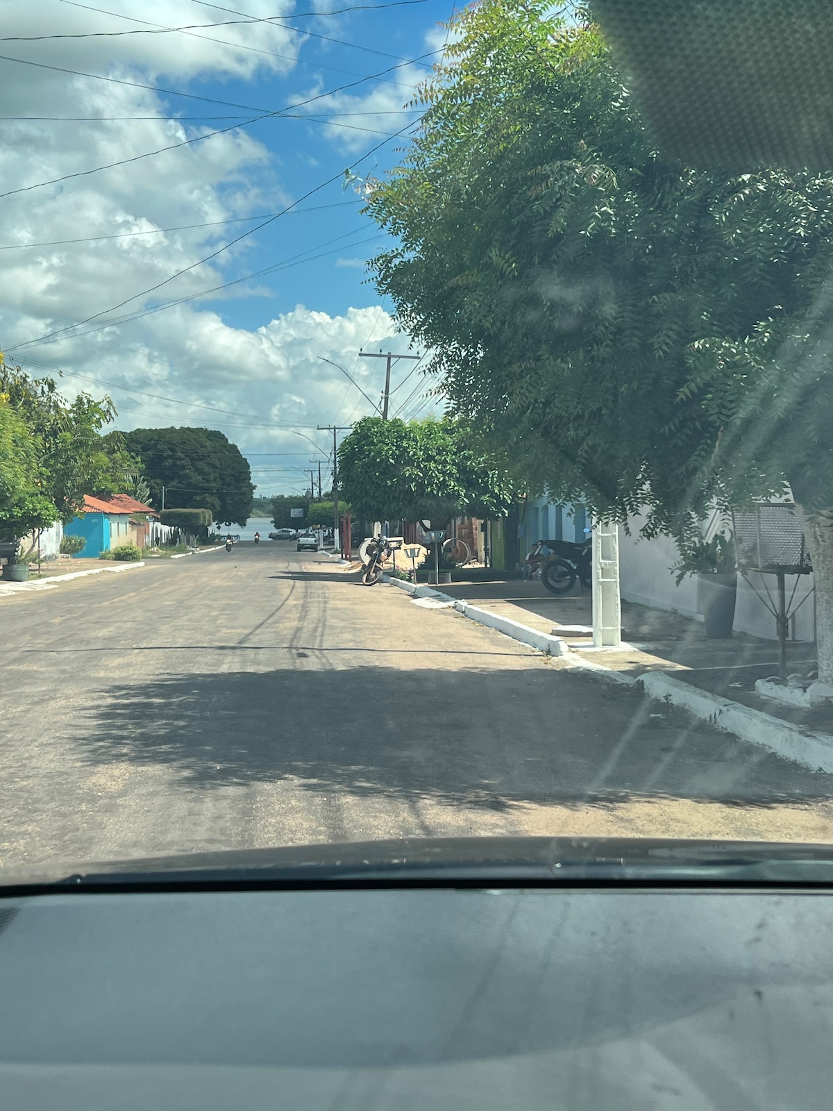 A neighborhood street is shown, lined with trees providing patches of shade. Colorful houses are visible along the side, with bicycles parked and subtle details of everyday life seen in the background. The sky is partly cloudy with blue and white hues.