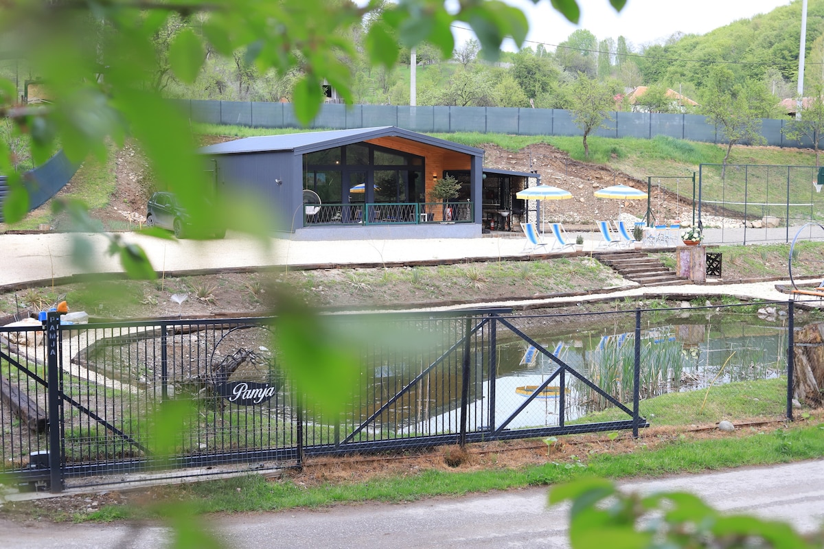 A modern building is viewed through lush greenery, surrounded by a tranquil pond. Leisure chairs and umbrellas sit nearby, highlighting outdoor relaxation areas. A fenced pathway leads to the entrance, suggesting accessibility to various recreational opportunities.