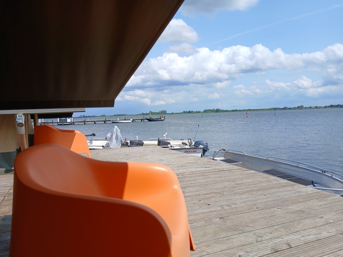 A wooden deck extends towards the water, with comfortable orange chairs positioned for relaxed viewing. Boats are anchored nearby, and gentle waves reflect the sky. The horizon features green land and a few structures, complemented by a light cloud cover.