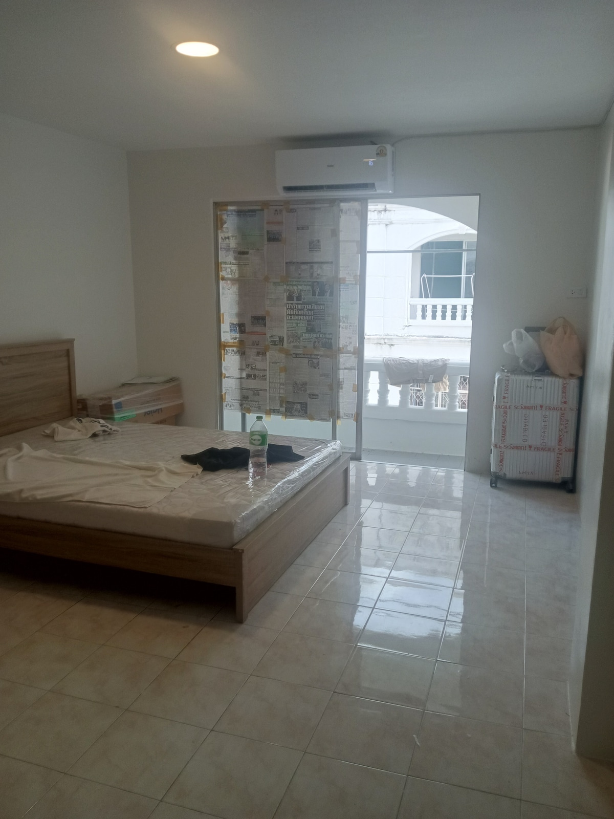 A bedroom features a wooden bed frame with unmade bedding and a wall-mounted air conditioning unit. Natural light enters through a glass door leading to a small balcony, while a storage chest and a stuffed animal are positioned nearby on the floor.