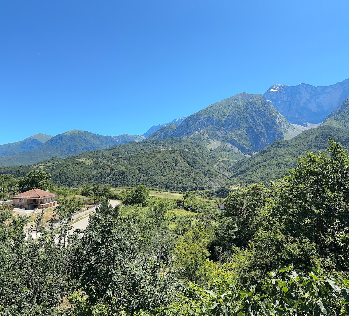 A serene landscape showcases rolling green hills and a clear blue sky. In the foreground, lush greenery frames a winding road. A distant mountain range stands majestically, adding depth to the tranquil setting. A single building is visible in a valley, surrounded by nature.