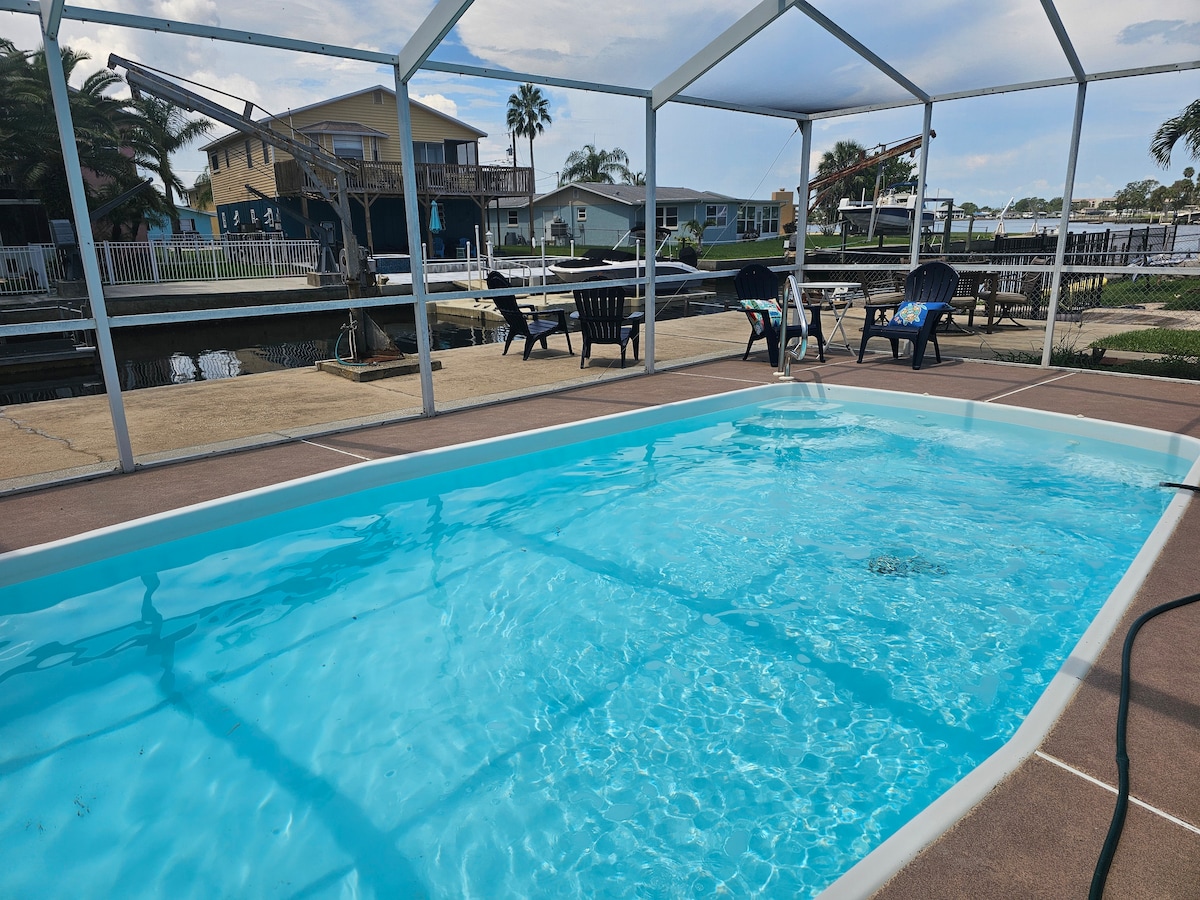 A private pool is positioned within a screened enclosure, featuring clear blue water. Sun loungers and dining tables are arranged nearby, with views of the canal and surrounding palm trees visible in the background.