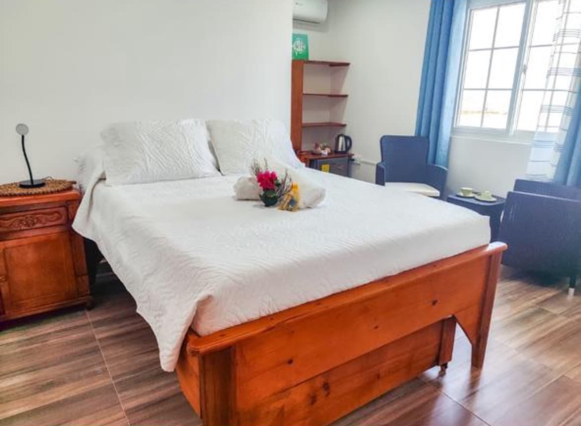 A spacious bed is dressed in white linens, with decorative towels and a small flower arrangement on top. Nearby, a wooden nightstand and a seating area with two chairs are visible. Light enters through the window, complemented by curtains in a soft blue.