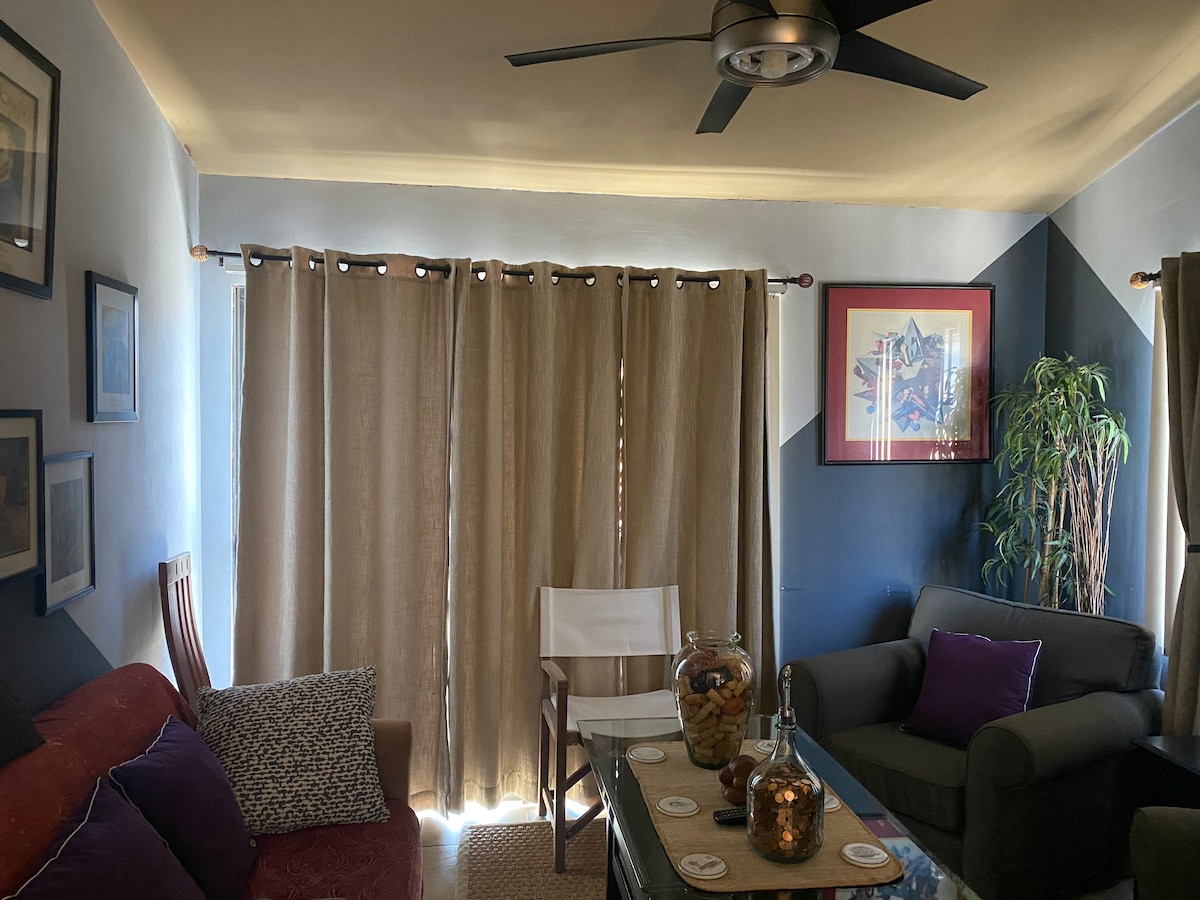 A cozy living area features a comfortable sofa adorned with purple pillows and a green armchair. Natural light is filtered through beige curtains, and artwork is displayed on the walls. A decorative table holds a glass jar filled with colored stones and a collection of coasters.
