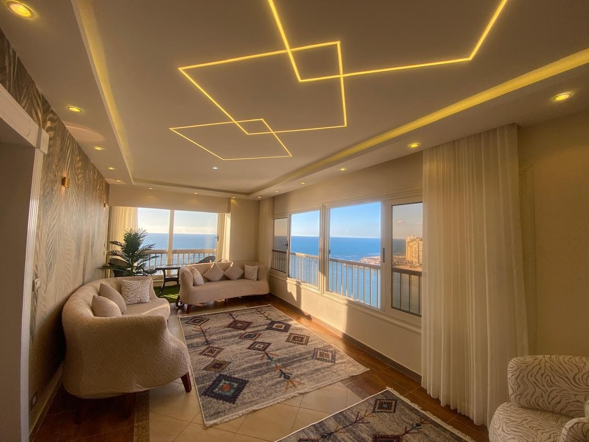A bright living area features panoramic windows that provide sweeping views of the sea. Soft seating arrangements are complemented by a decorative area rug that adds texture to the space. Ambient lighting from the ceiling enhances the room's bright and airy feel.