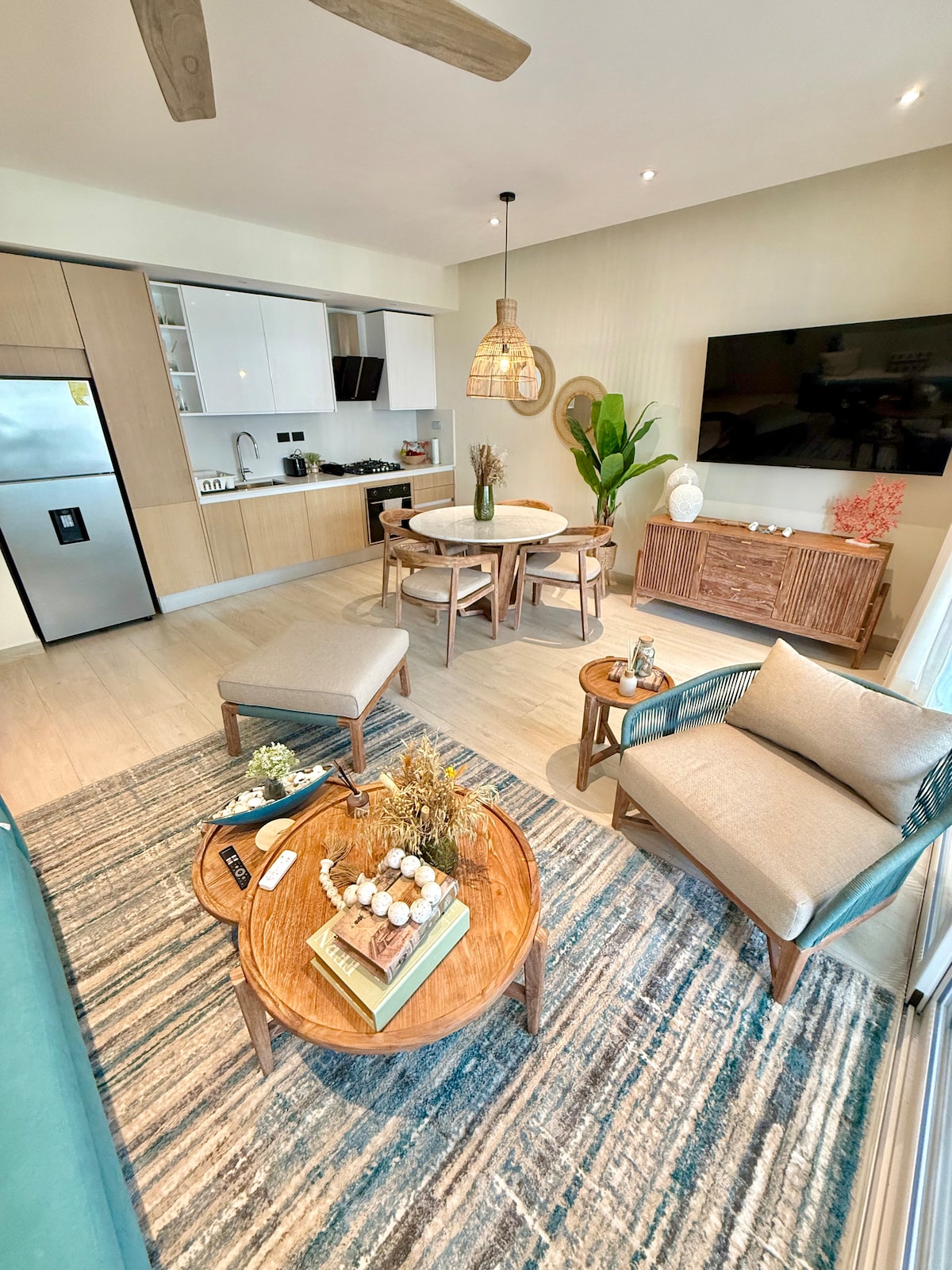 The living area is characterized by wooden furniture, including a round dining table with chairs and two cozy armchairs. A large flat-screen television is mounted on the wall, while indoor plants and decorative items add a touch of nature to the space.