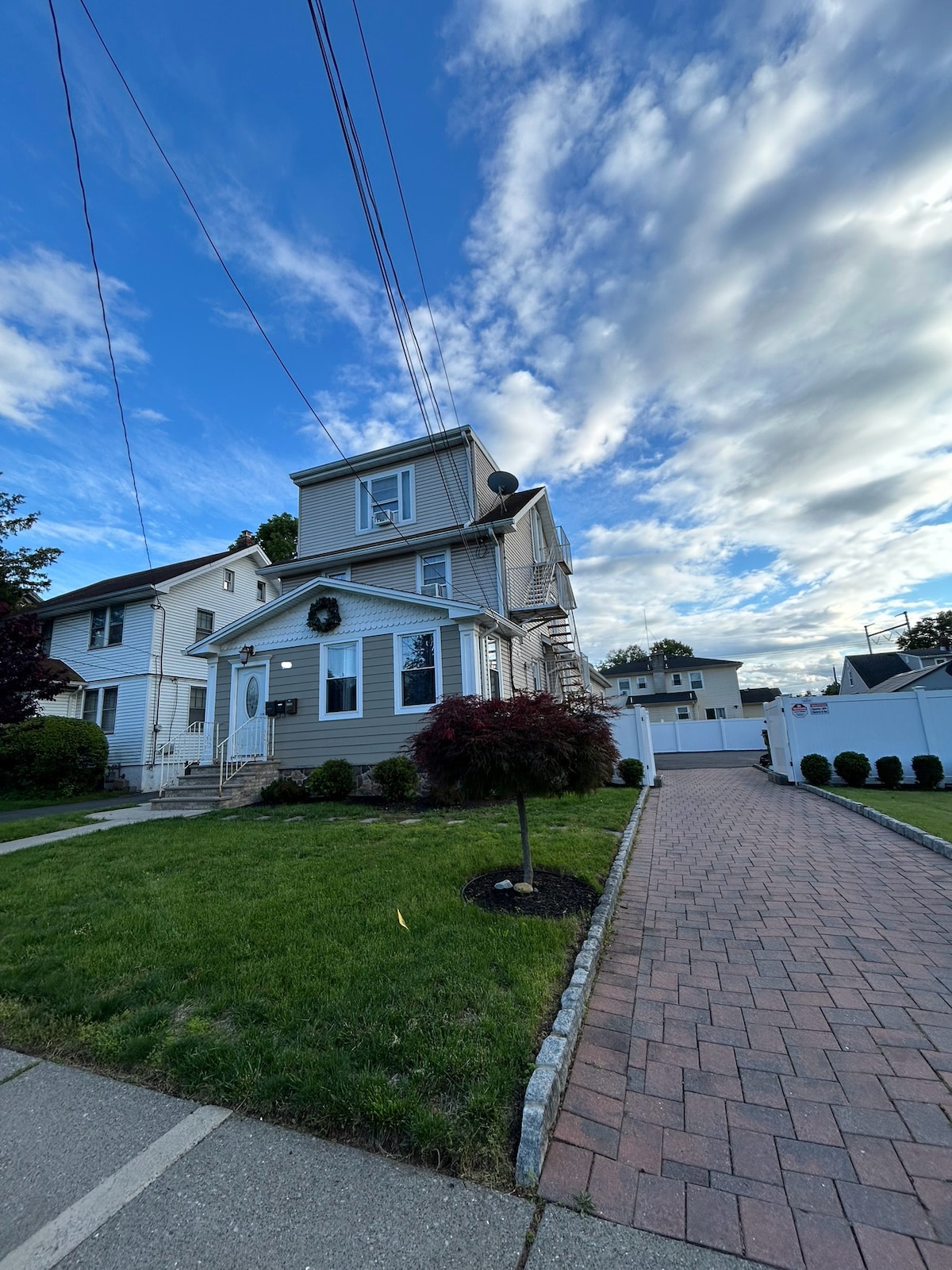 A well-maintained two-story building is depicted, featuring beige siding and a welcoming porch. The front yard is landscaped with neatly trimmed shrubs, and a paved driveway leads to a gated area. A clear blue sky with wispy clouds enhances the serene setting.