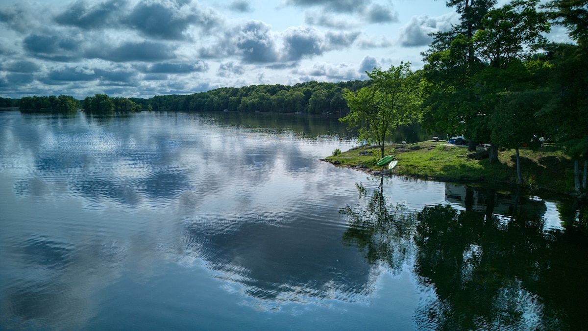 A serene lakeside view features calm water reflecting the sky and surrounding greenery. Lush trees and a small shoreline are visible, providing a tranquil space for relaxation. Kayaks are positioned near the water's edge, suggesting opportunities for outdoor activities.