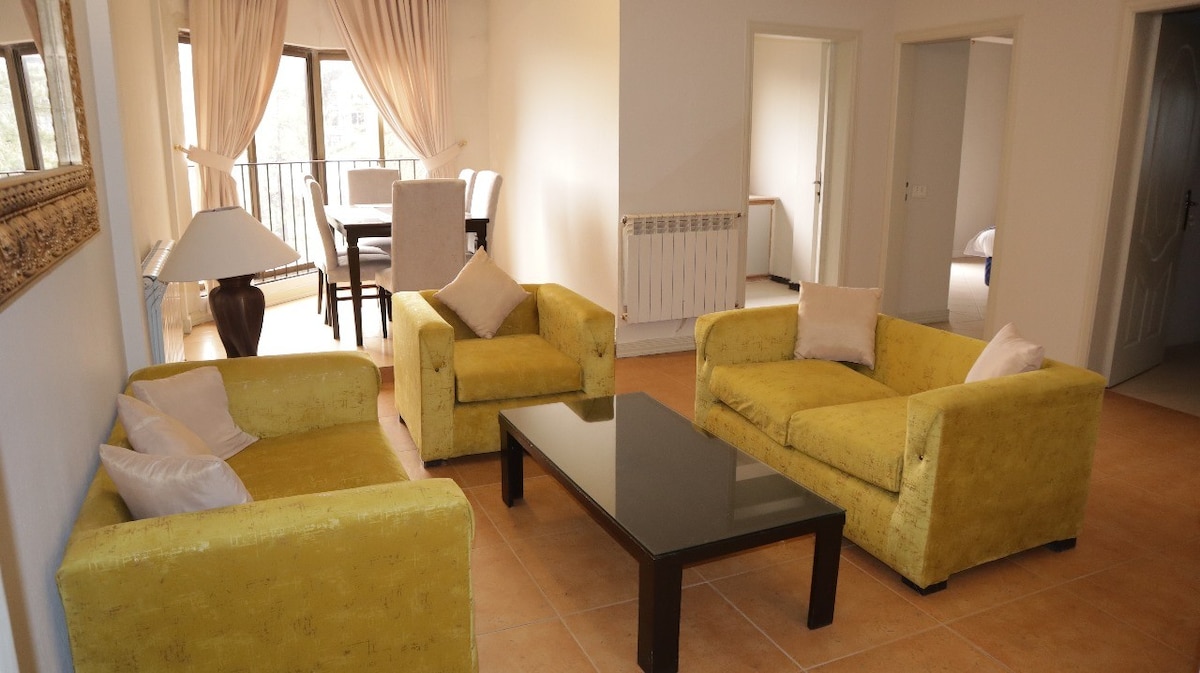 A spacious living area features two bright yellow sofas with white cushions, arranged around a dark coffee table. Natural light fills the room through large windows with sheer curtains. A dining set is visible in the background, enhancing the open and welcoming layout.