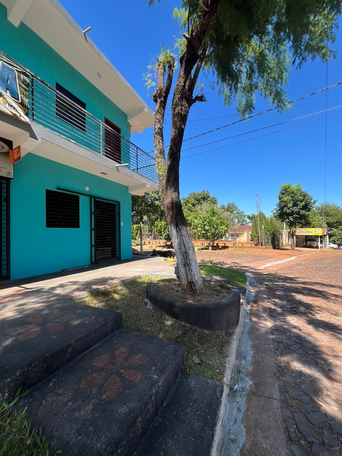 The exterior of a two-story building is shown, painted in a vibrant blue hue. A small tree stands nearby, with concrete steps leading to the entrance. The street is lined with greenery, and clear blue skies are visible overhead.