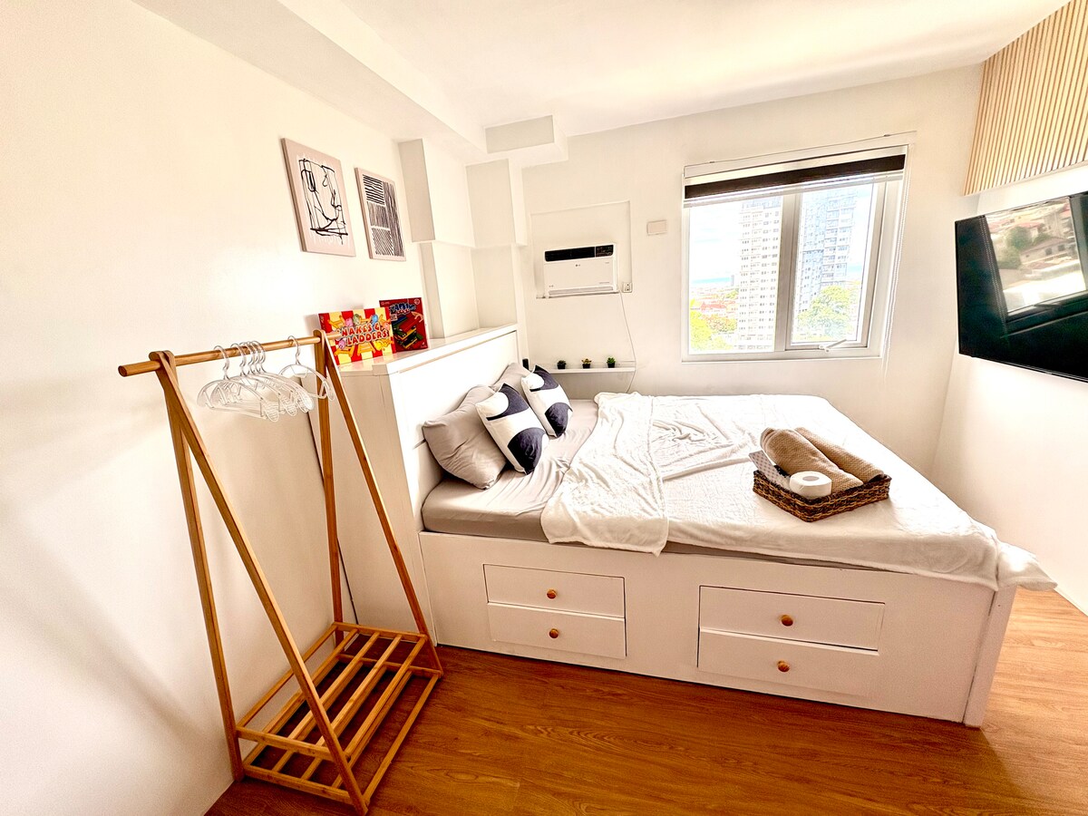 A well-lit bedroom features a spacious bed adorned with neutral-toned pillows and a cozy blanket. A clothing rack stands beside the bed, while a window offers views of the skyline. Minimalist decor emphasizes a clean, serene environment.