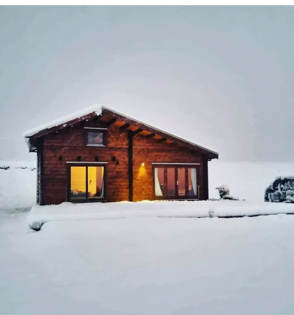 A rustic cabin is set against a snowy landscape, featuring wooden exterior and a sloped roof. Large windows reflect warm light from inside, while the surrounding area is covered in fresh snow. A cozy atmosphere is suggested by the snow-dusted surroundings.