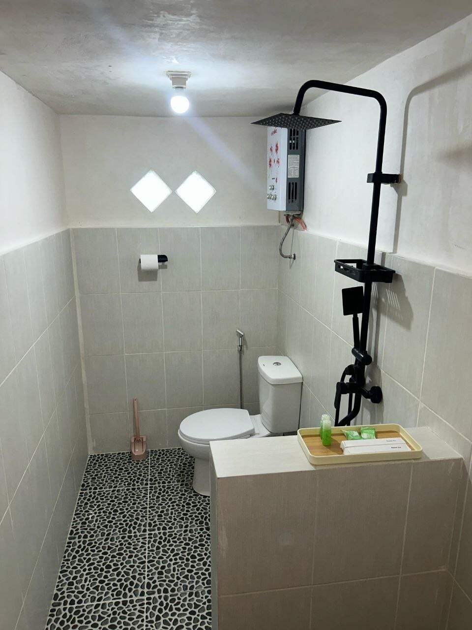 A modern bathroom is showcased, featuring a compact toilet and a shower with an overhead rain showerhead. The walls are tiled in neutral tones, and a small tray holds toiletries. A diamond-shaped window allows natural light to enter, creating an open feel.