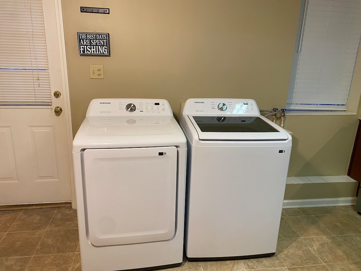 A laundry area is equipped with a stacked washer and dryer, featuring a clean white finish. Natural light enters through a nearby window fitted with horizontal blinds. A door leading outside is visible on the left side of the image.