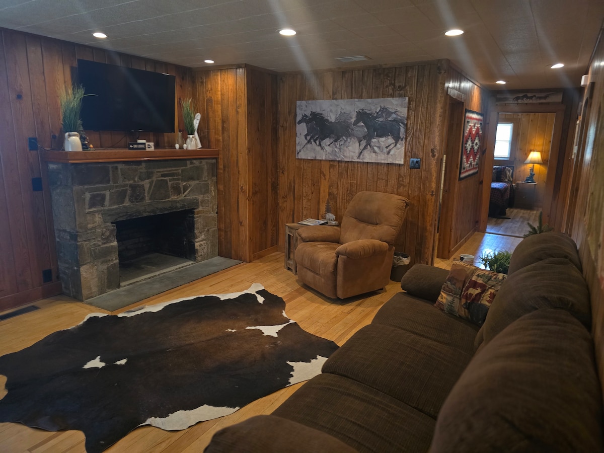 A comfortable living area features wood-paneled walls and warm lighting. A brown sectional sofa is situated in front of a stone fireplace, while a wall-mounted television is positioned above the mantel. Western-themed artwork enhances the decor, complemented by a cozy armchair.