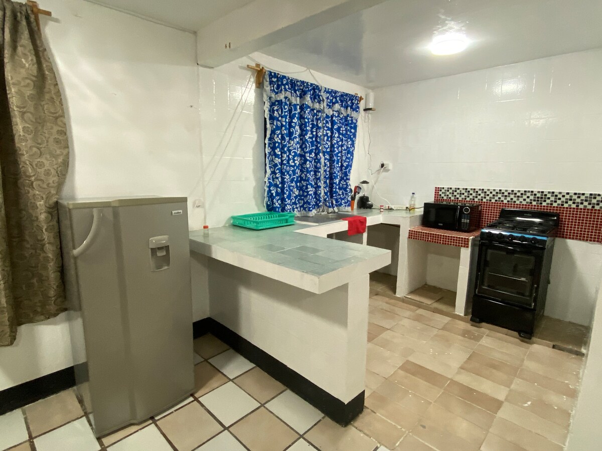 The kitchen features a refrigerator, a microwave, and a stove with an oven. Counter space is provided for food preparation, alongside a sink. Bright blue curtains frame a window, allowing natural light to enter the room. The tiled floor and wall accents add a practical touch.